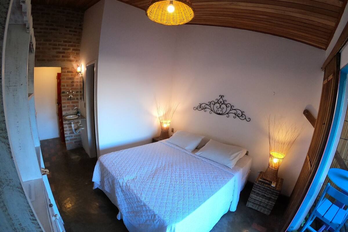 Quarto com cama, duas luminárias de cabeceira, um objeto decorativo na parede e uma cesta de vime.