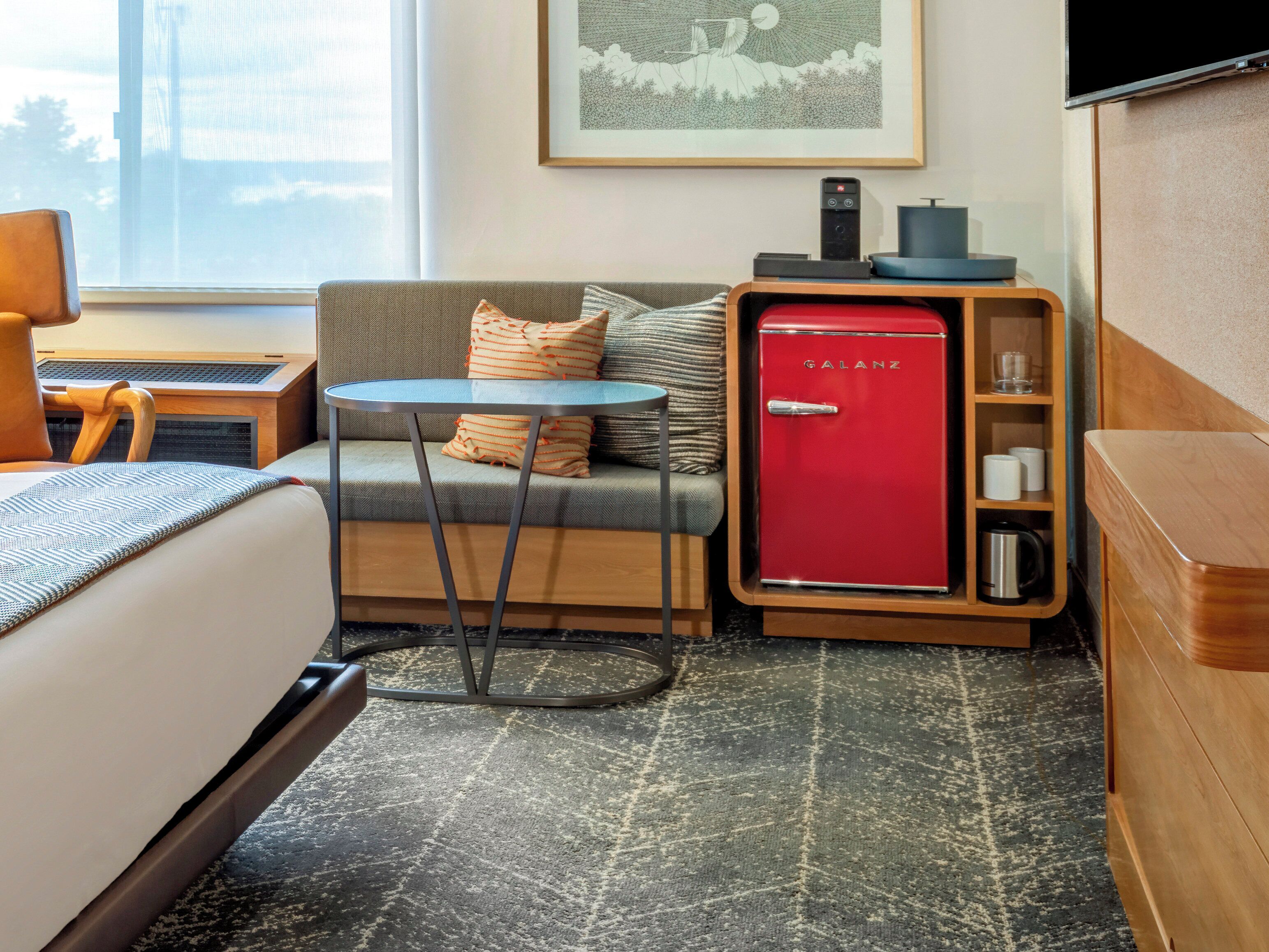 Una habitación de hotel con cama, sofá, una mesita, un refrigerador Galanz rojo, un armario de madera y un cuadro en la pared.