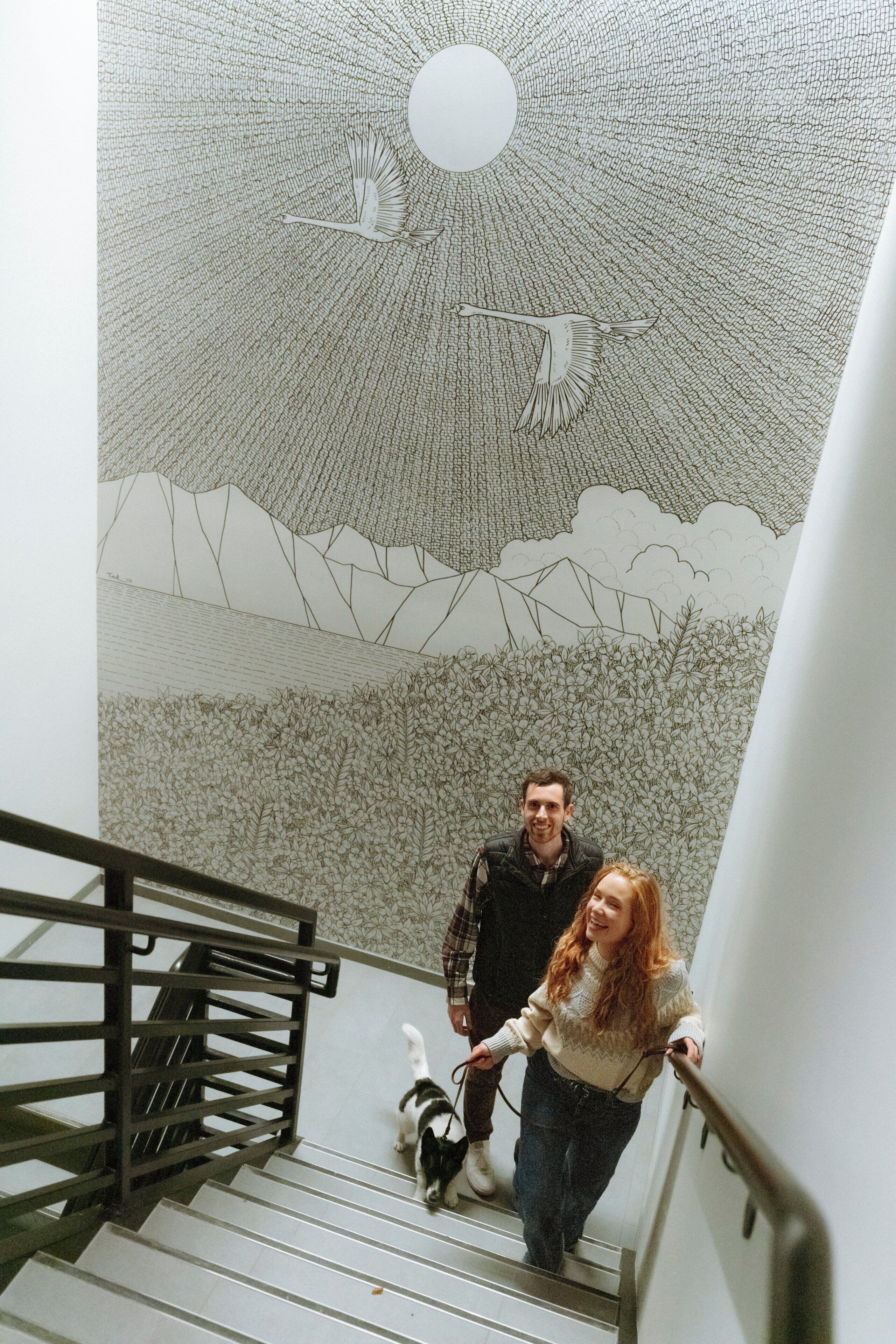 A staircase with a detailed mural on the wall, a metal handrail, and a person walking a dog.
