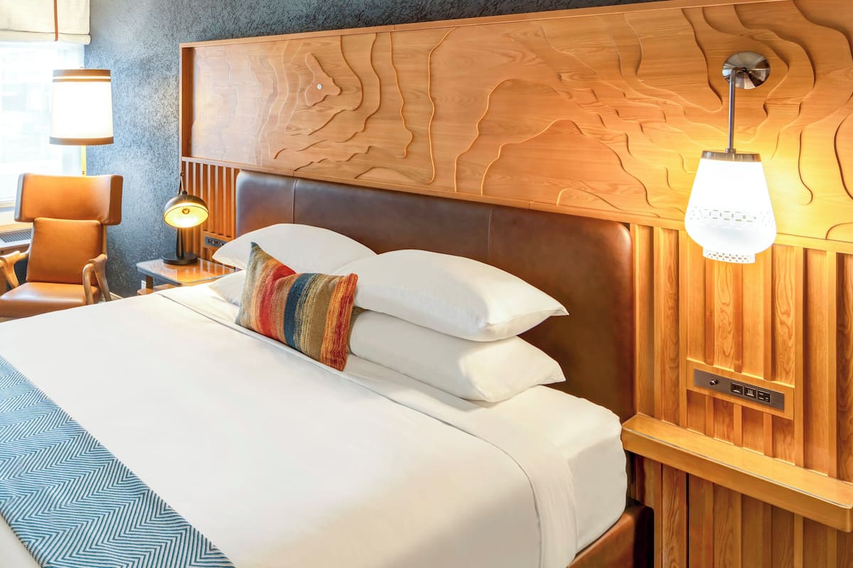 A hotel room with a wooden headboard, a bedside table with a lamp, a bed with white linens and a colorful pillow, and a wall-mounted light fixture.