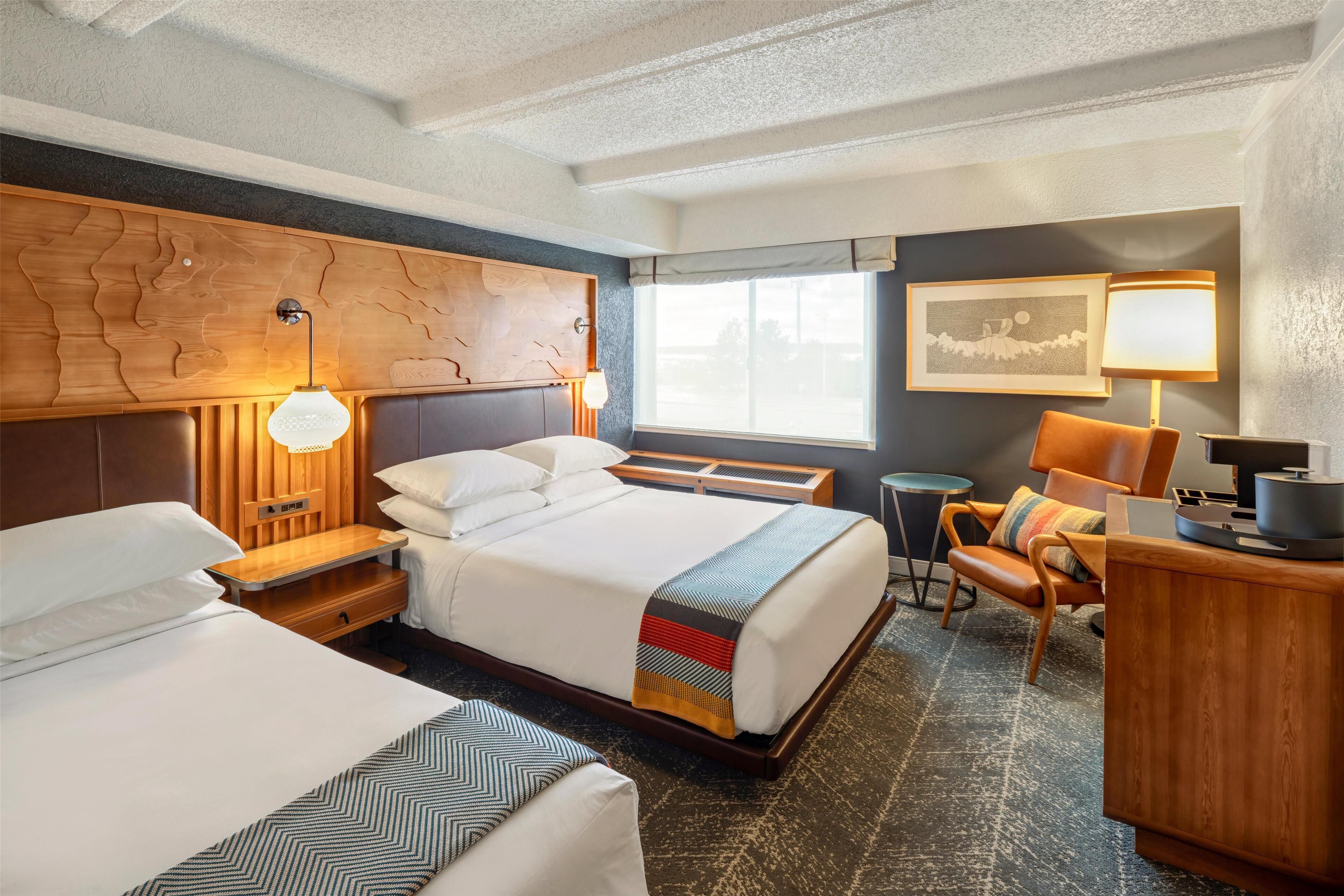 A hotel room with two beds, a wooden headboard, a desk with a lamp, a chair, and a window with a view.
