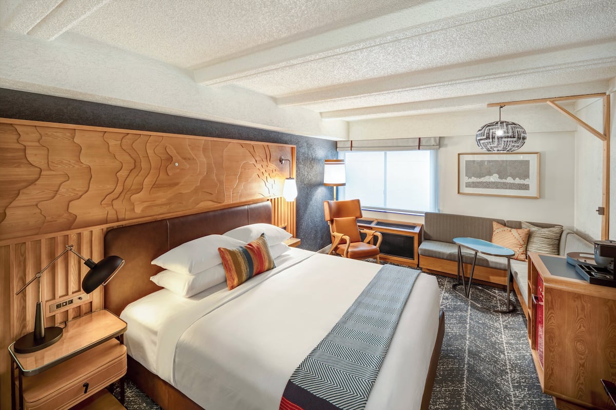 A hotel room with a large bed, a wooden headboard, a desk with a lamp, a chair, a sofa, and a window with a view.