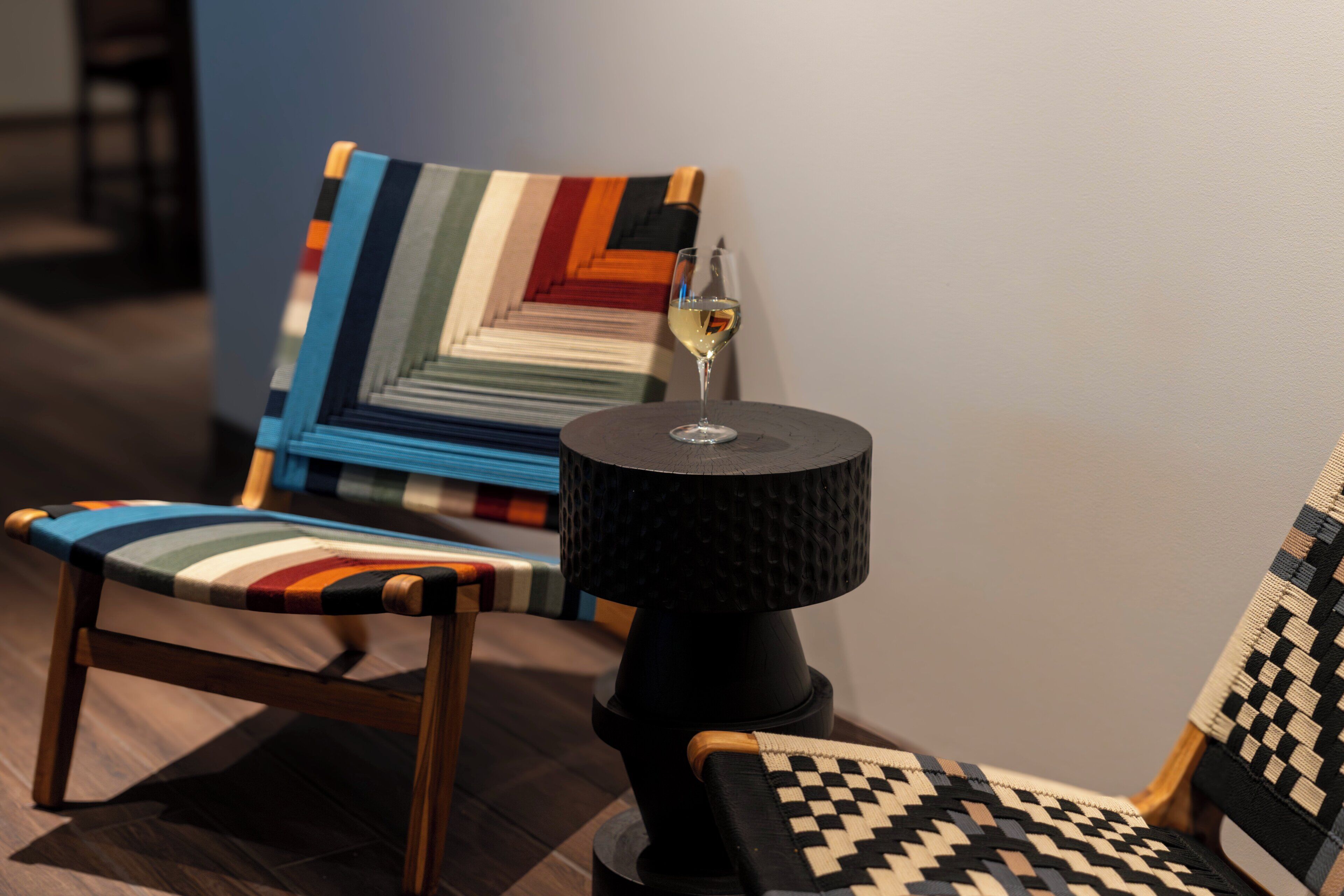 Quarto moderno com cadeira de madeira, poltrona estampada e pequena mesa com um copo de vinho.