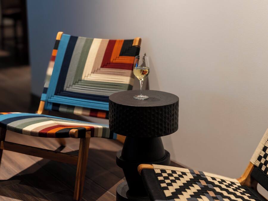 Una habitación moderna con una silla de madera, un sillón estampado y una mesita con una copa de vino.