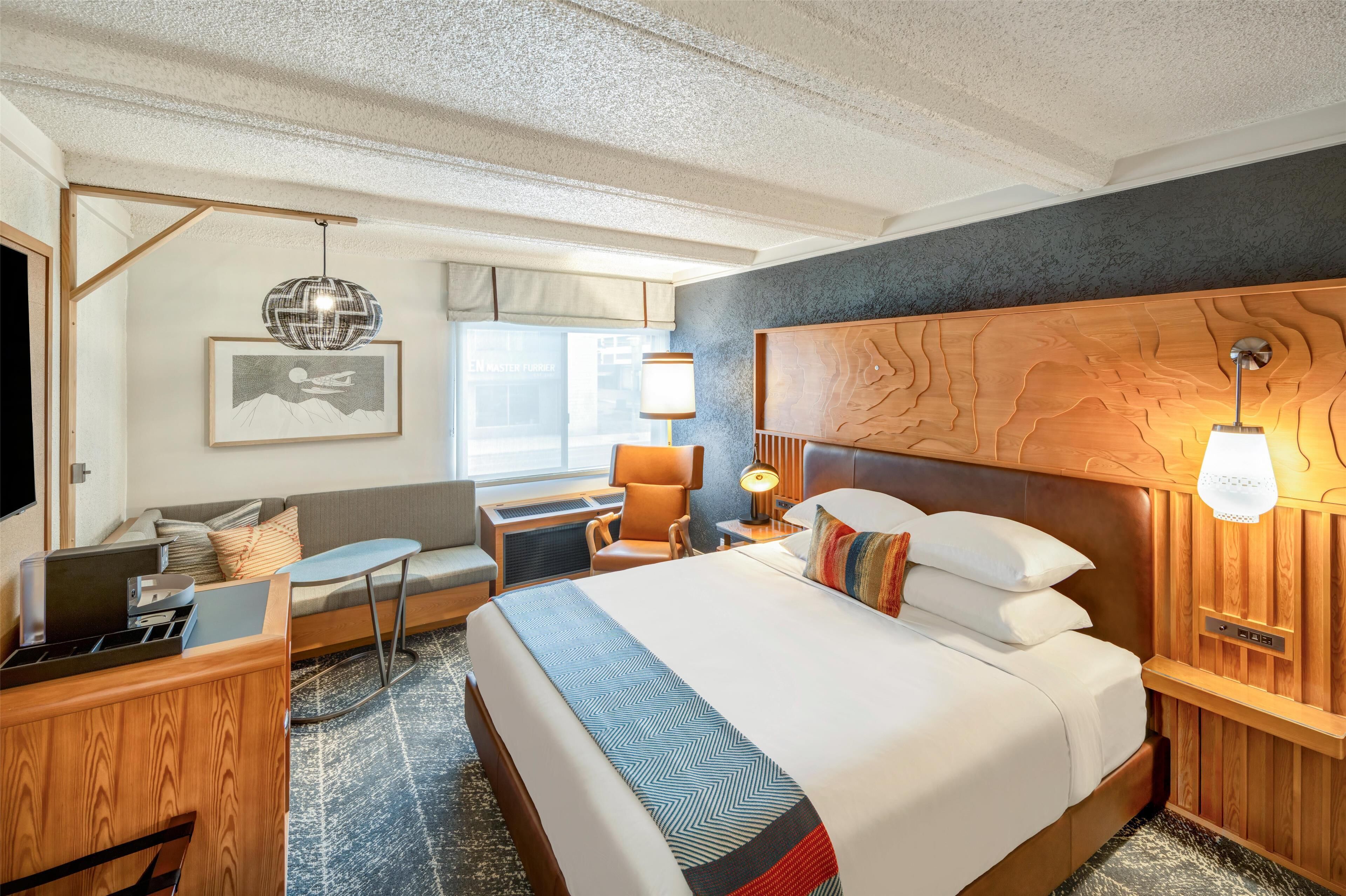 A hotel room with a large bed, a desk with a laptop, a chair, a lamp, and a wall-mounted TV.