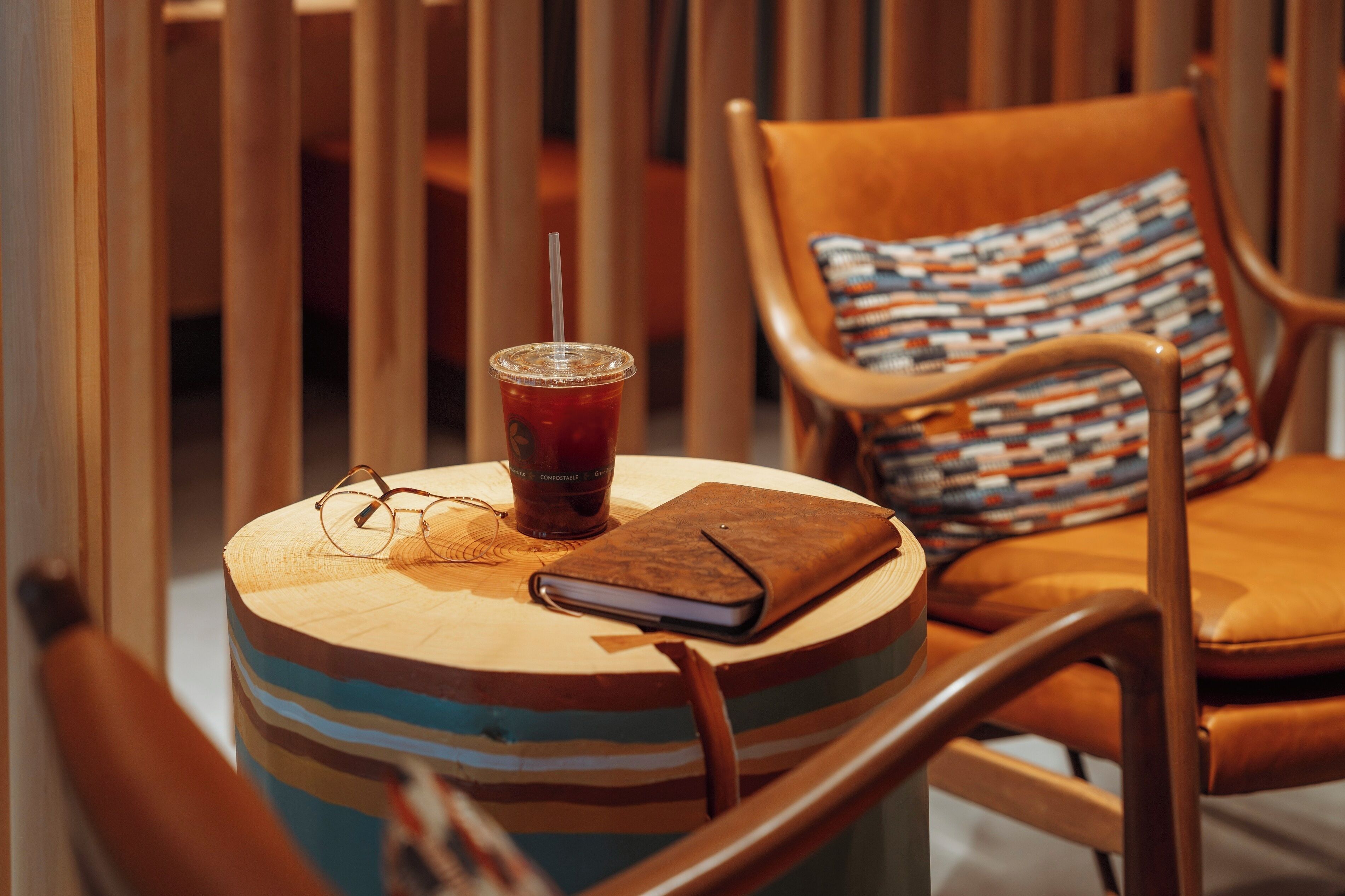 A round wooden table with a striped cushion, a drink with a straw, and a closed notebook.
