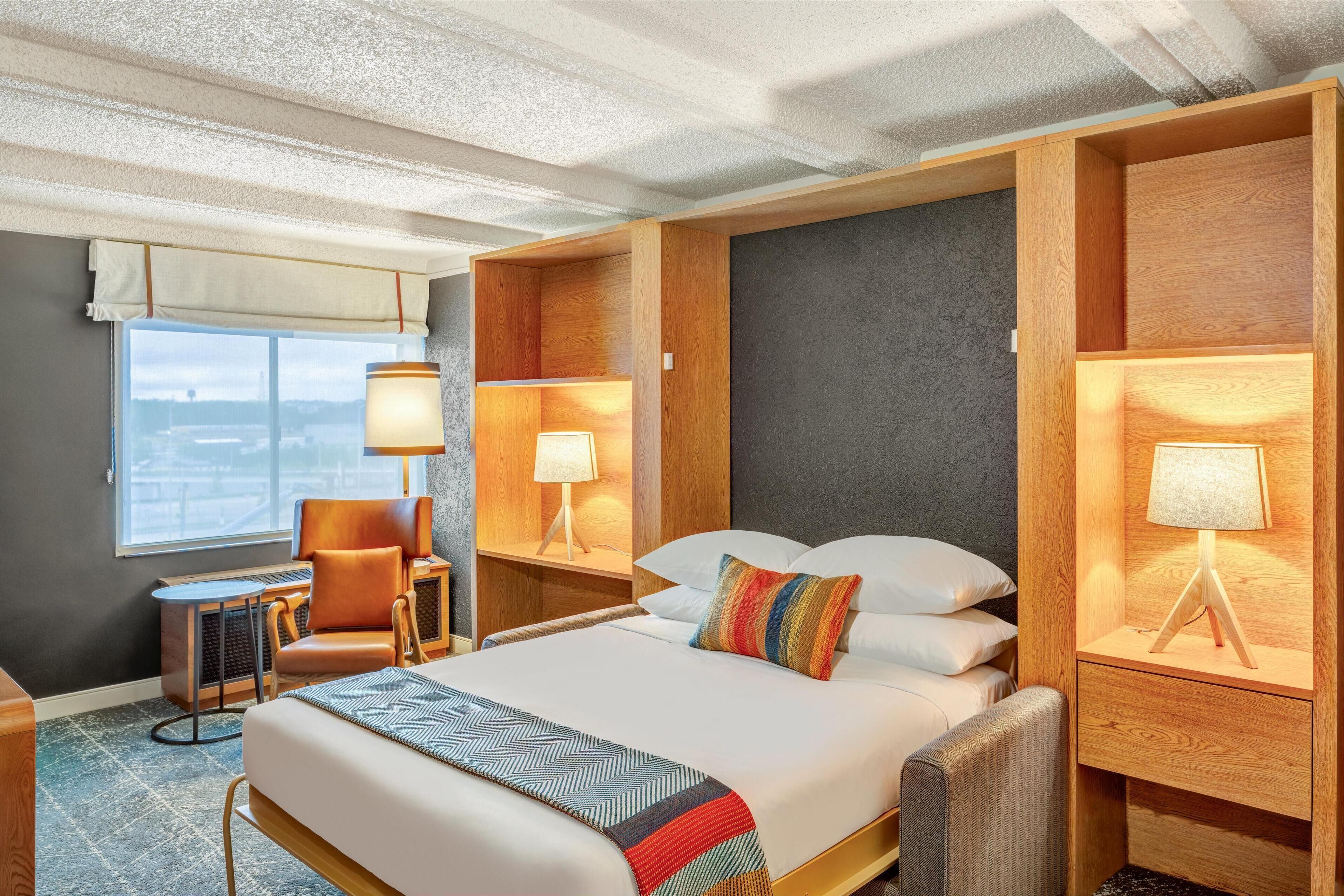 A hotel room with a large bed, two bedside tables with lamps, a chair, and a view of the city through the window.