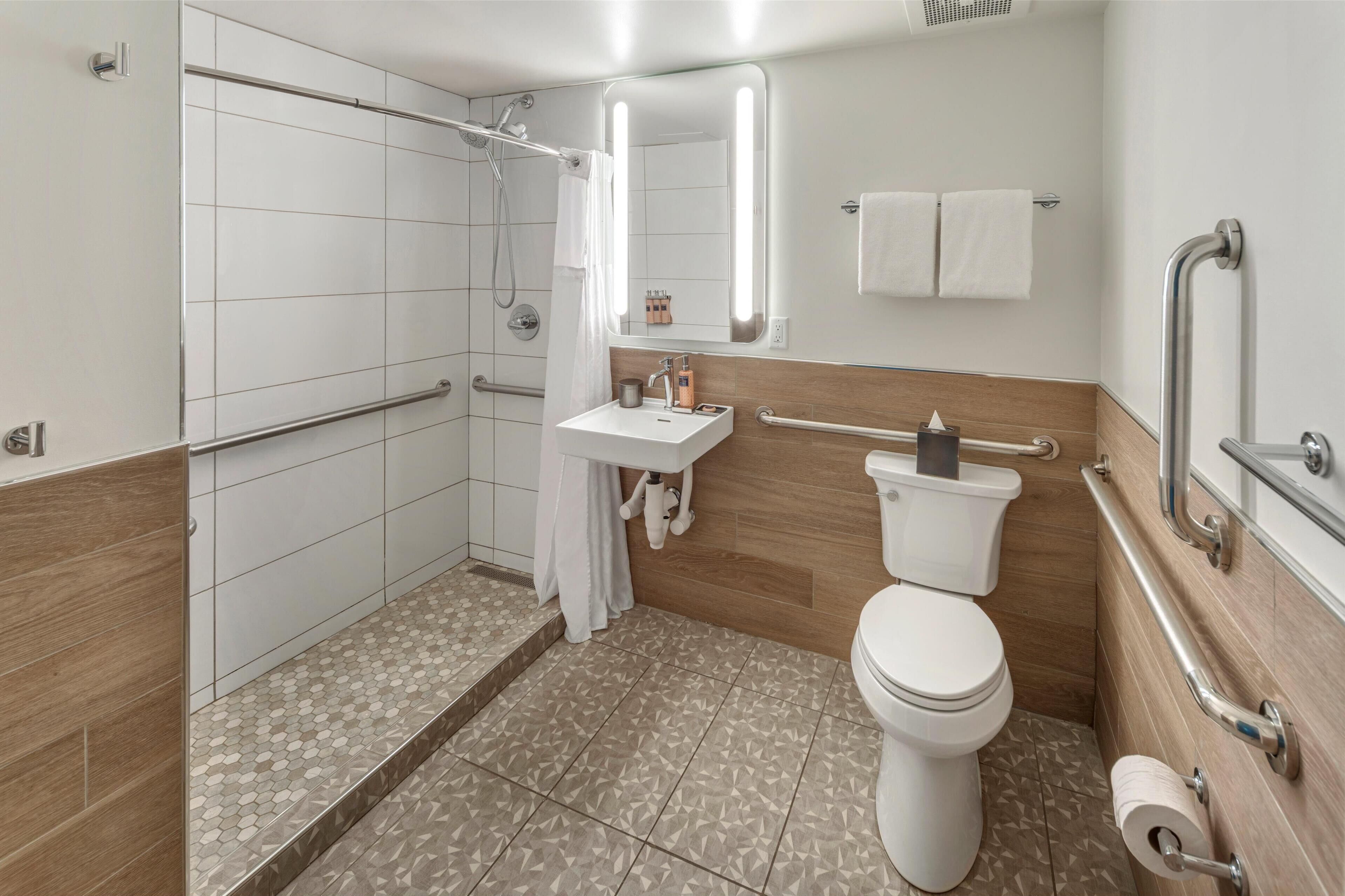 Een moderne badkamer met douche, toilet en wastafel. Er zijn handgrepen en een handrail voor toegankelijkheid.