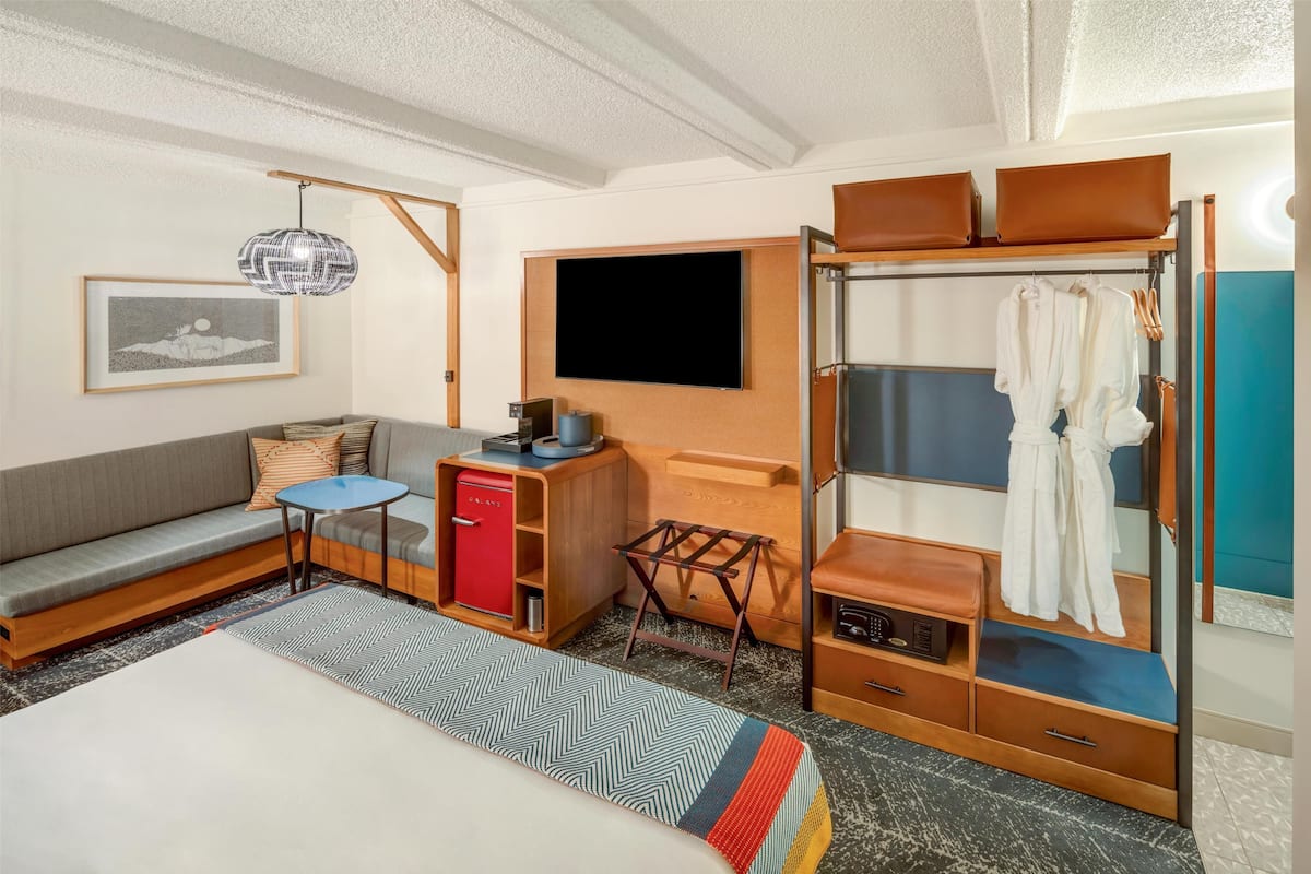 A compact hotel room with a sofa, a small table, a TV, a wardrobe, and a bed.