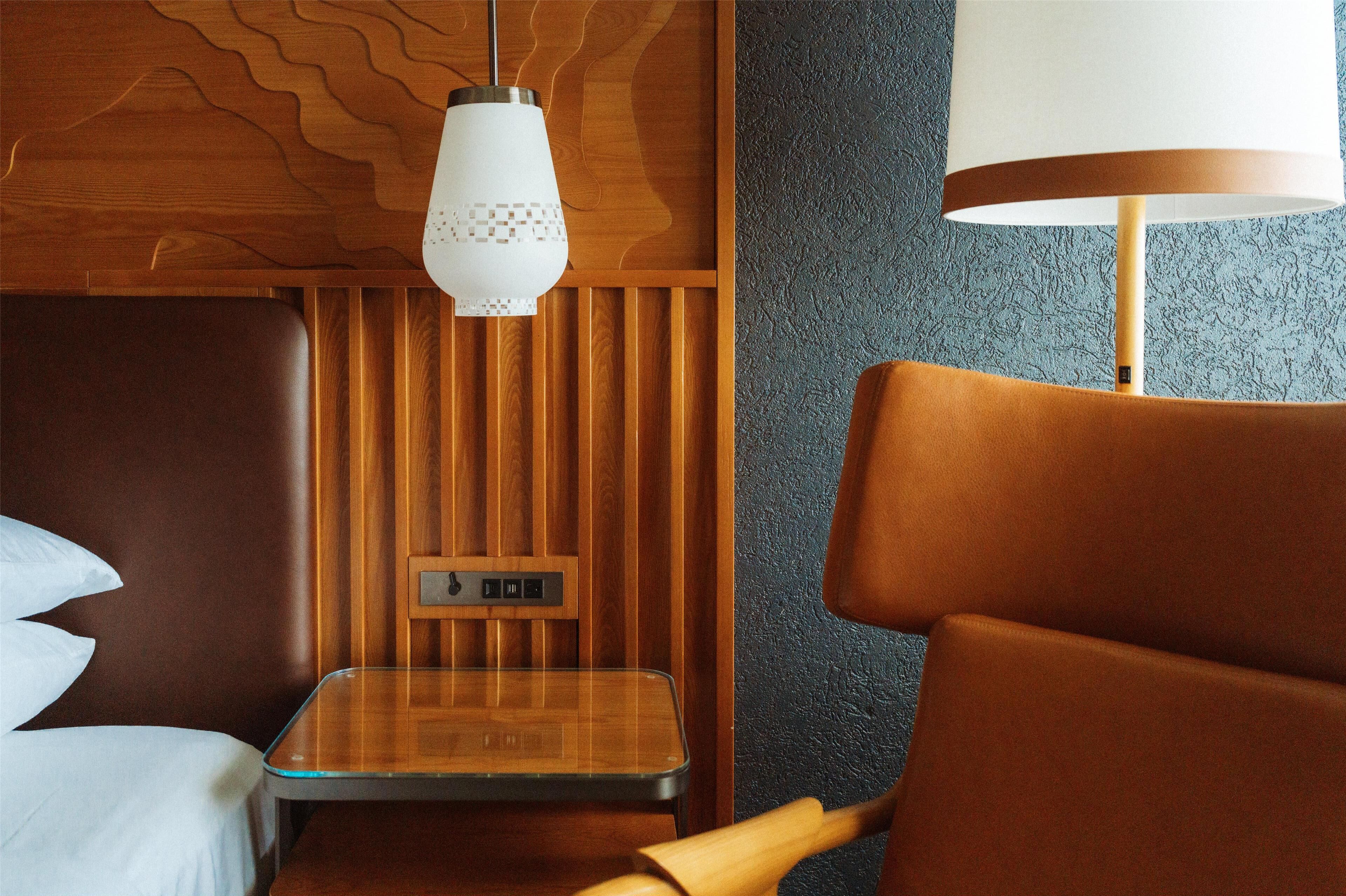 A hotel room with a wooden headboard, a small wooden table, a modern lamp, and a control panel on the wall.