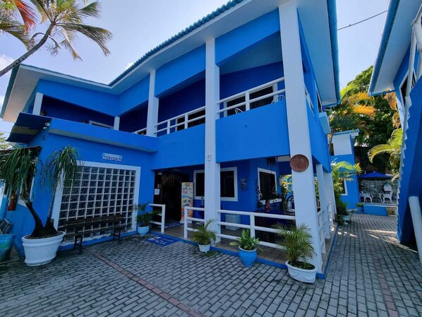 A two-story building with a blue and white color scheme, featuring a reception area with a sign, potted plants, and a tiled entrance. - Morada Azul (Ipojuca)