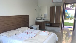 A neatly made bed with white linens and a quilt, a small bedside table with a bottle, a mirror above a wooden cabinet, and a view of a garden through an open door. - Morada Azul (Ipojuca)