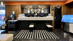 Een receptiegebied van een hotel met een modern design, met een receptiebalie, een kiosk voor digitaal inchecken en een groot scherm dat het interieur van het hotel toont.