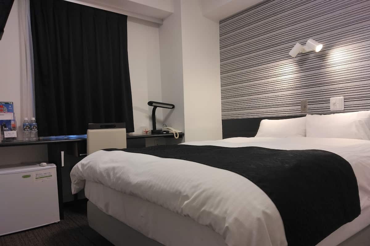 Une chambre d’hôtel avec un lit, un bureau avec une lampe, une chaise, un petit réfrigérateur et une fenêtre avec des rideaux.