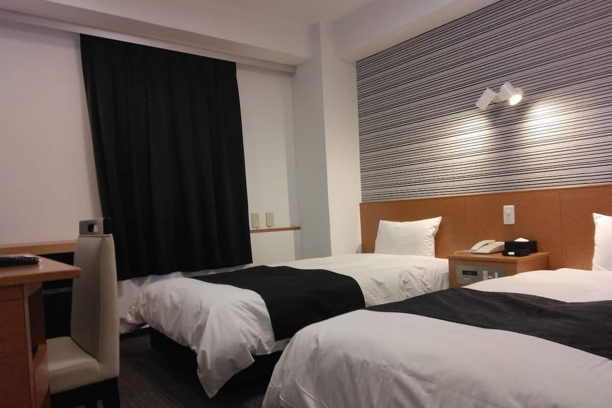 Une chambre d’hôtel avec deux lits, un bureau, une chaise, un téléphone et un luminaire fixé au mur.