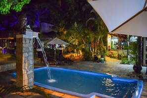A swimming pool with a waterfall feature, surrounded by lounge chairs and umbrellas, set in a landscaped garden with tropical plants. - Pousada Recanto da Coruja (Ipojuca)