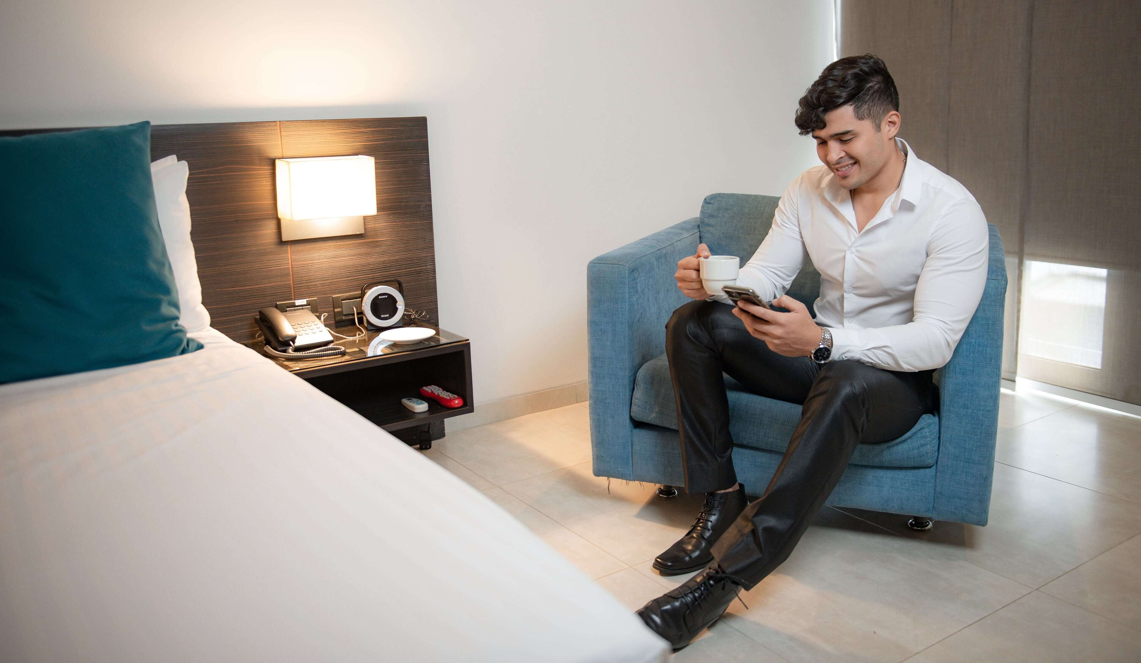 Una habitación de hotel moderna con una cama, un sillón azul, una mesita de noche con una lámpara, un teléfono y un control remoto.