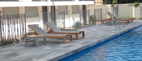 A poolside area with lounge chairs and umbrellas, a clear pool, and a modern building with balconies.