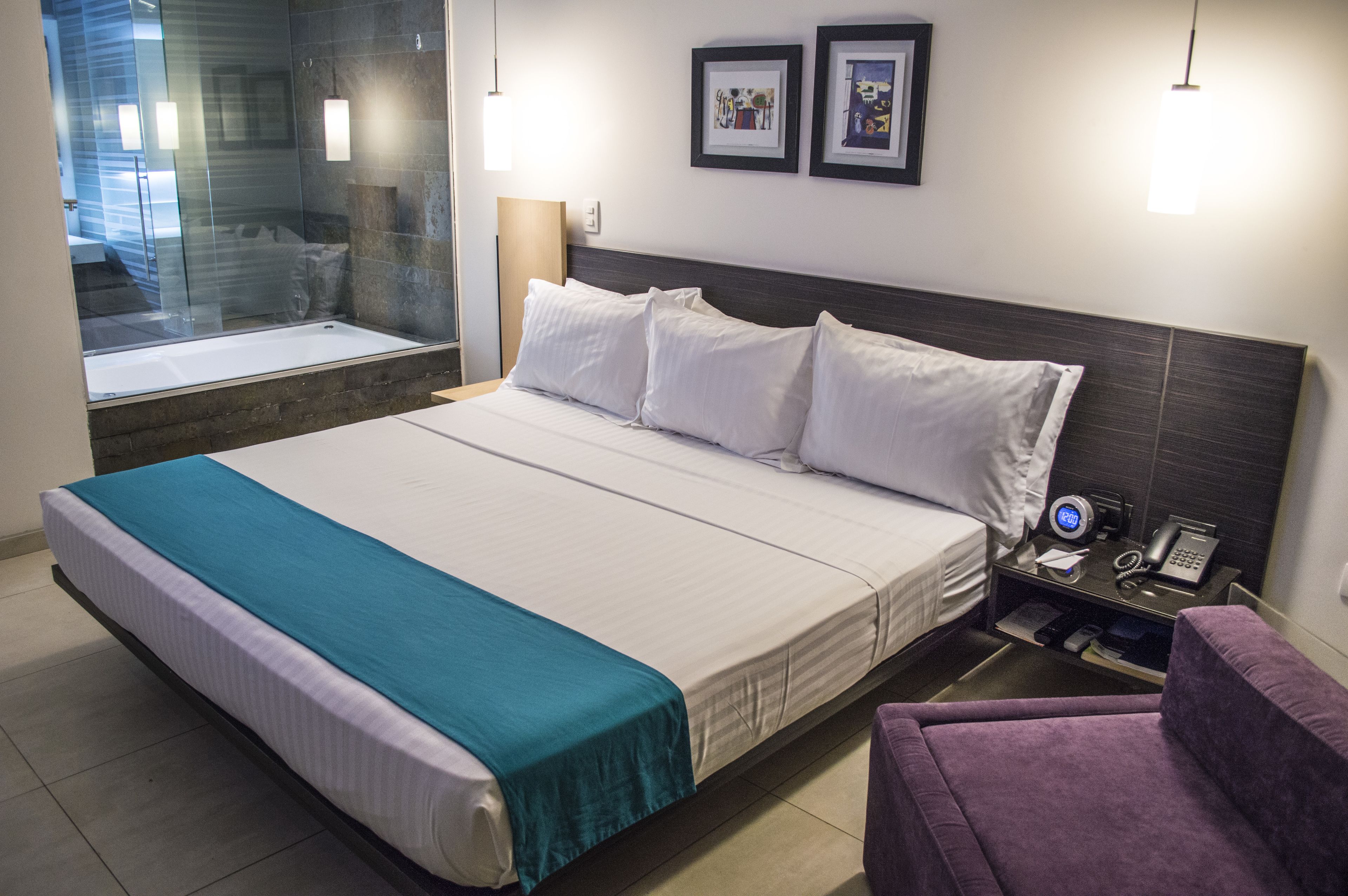 A hotel room with a large bed, a bedside table with a phone and a lamp, a purple armchair, and a view of a bathroom with a glass shower.