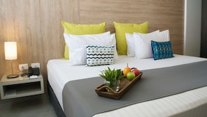 A neatly made bed with a grey bedspread, two yellow pillows, a blue and white patterned pillow, and a blue patterned pillow. There is a small bedside table with a lamp, a phone, and a basket containing a plant and fruits. - Hotel Casa Blanca (San José de Cúcuta)