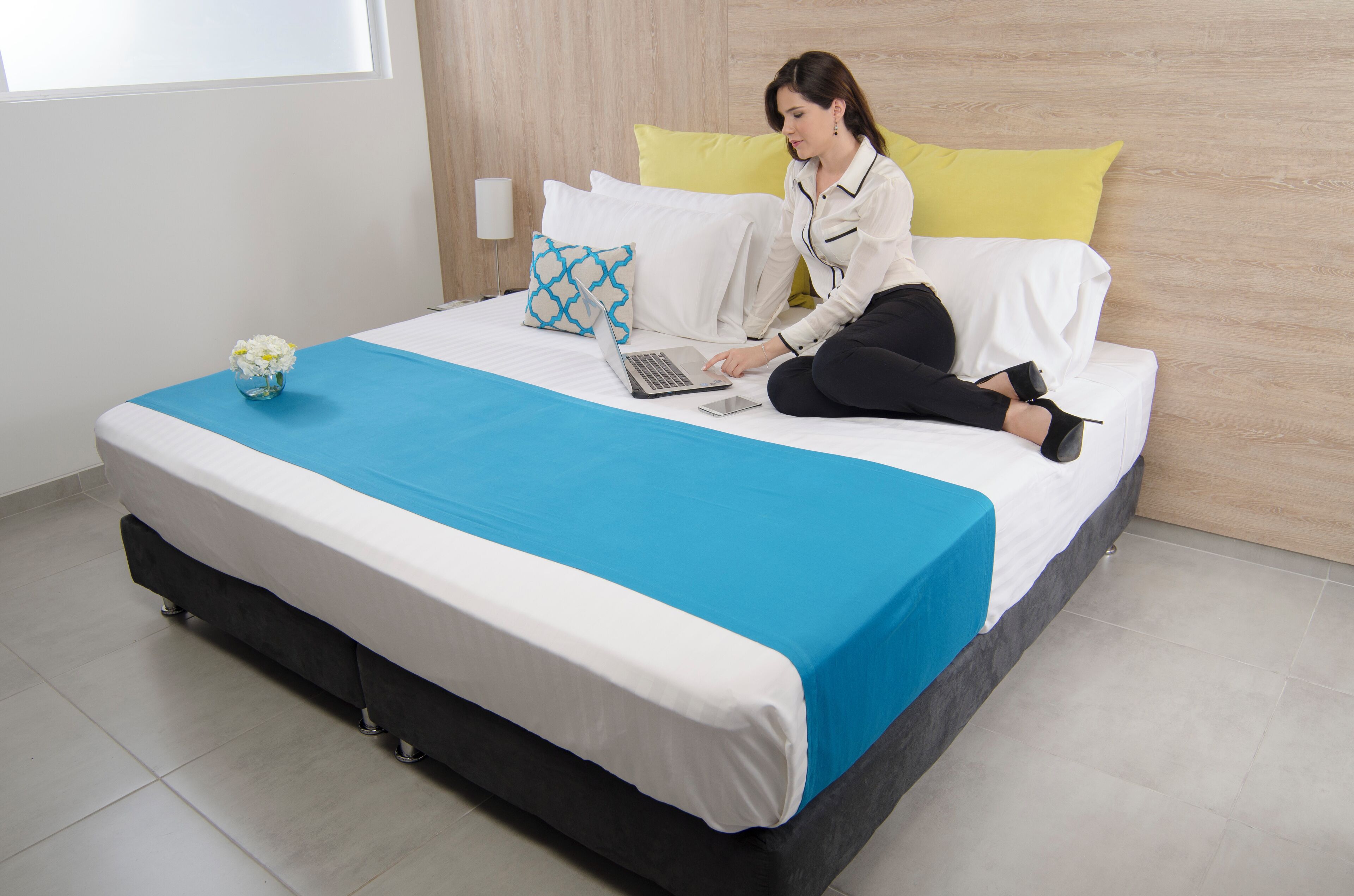 A bed with a laptop, a vase with flowers, and a yellow pillow.