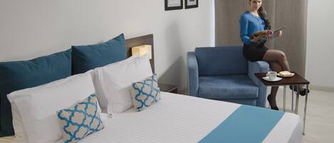 Habitación de hotel con una cama, un sillón azul, una mesita con una taza y un libro, y dos cuadros en la pared.