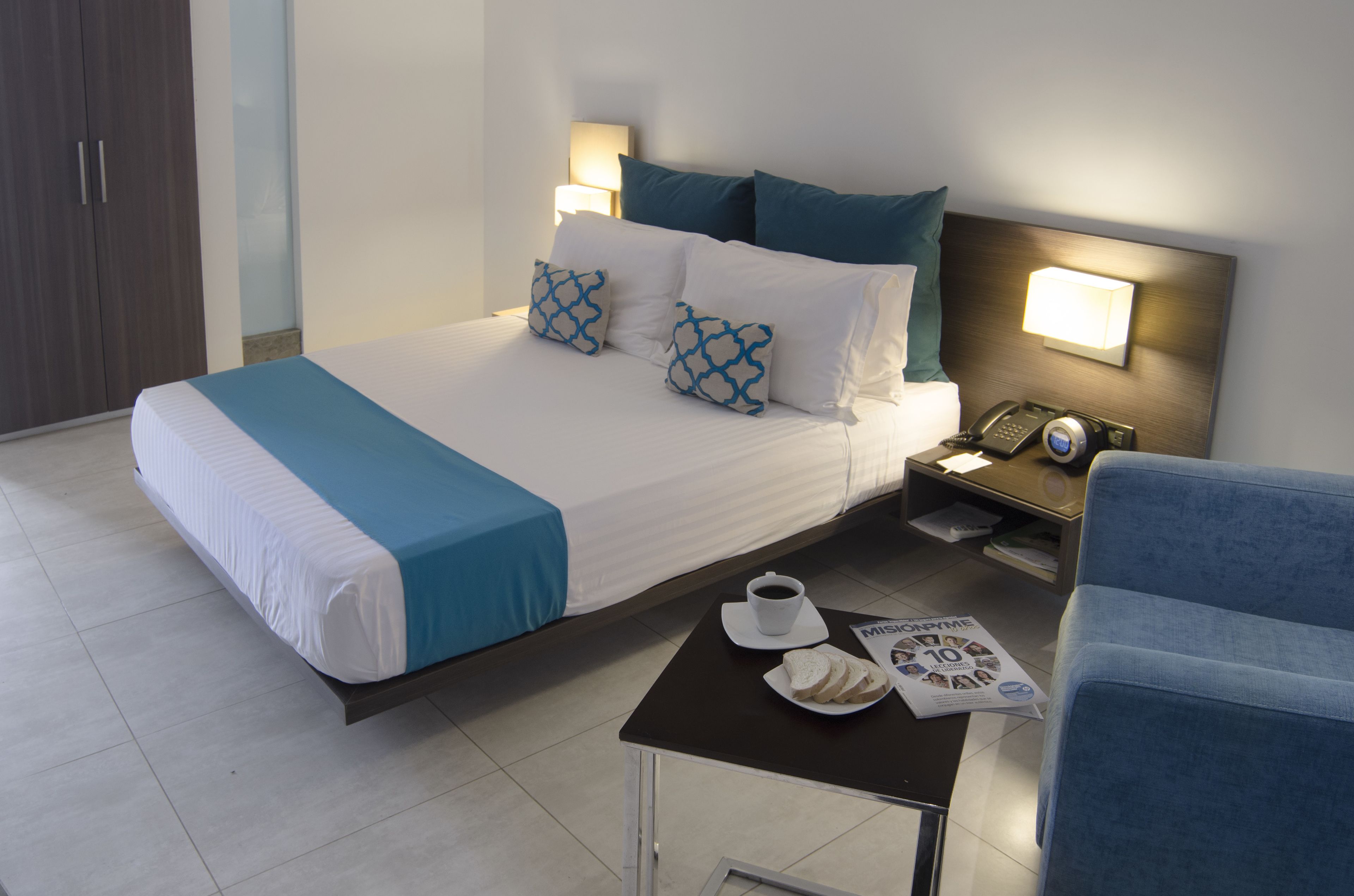 A hotel room with a bed, bedside table, chair, and a nightstand with a lamp and phone.