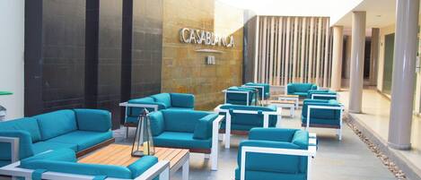 Un área de descanso moderna con sofás azul aguamarina y mesas blancas, una pared de piedra con el nombre "CASABLANCA" y un pasillo con columnas.