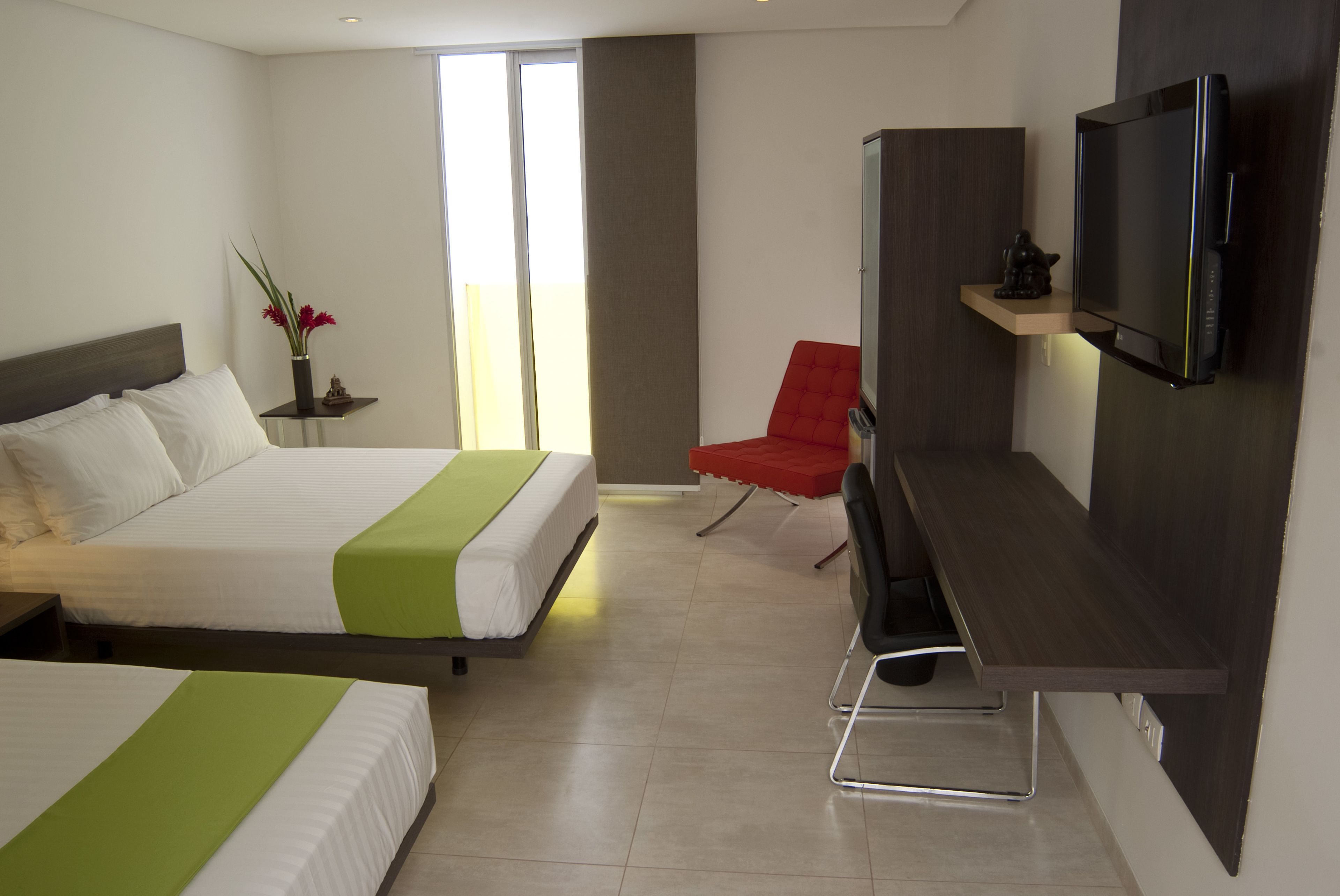A hotel room with two beds, a TV, a red chair, and a small table with a statue.