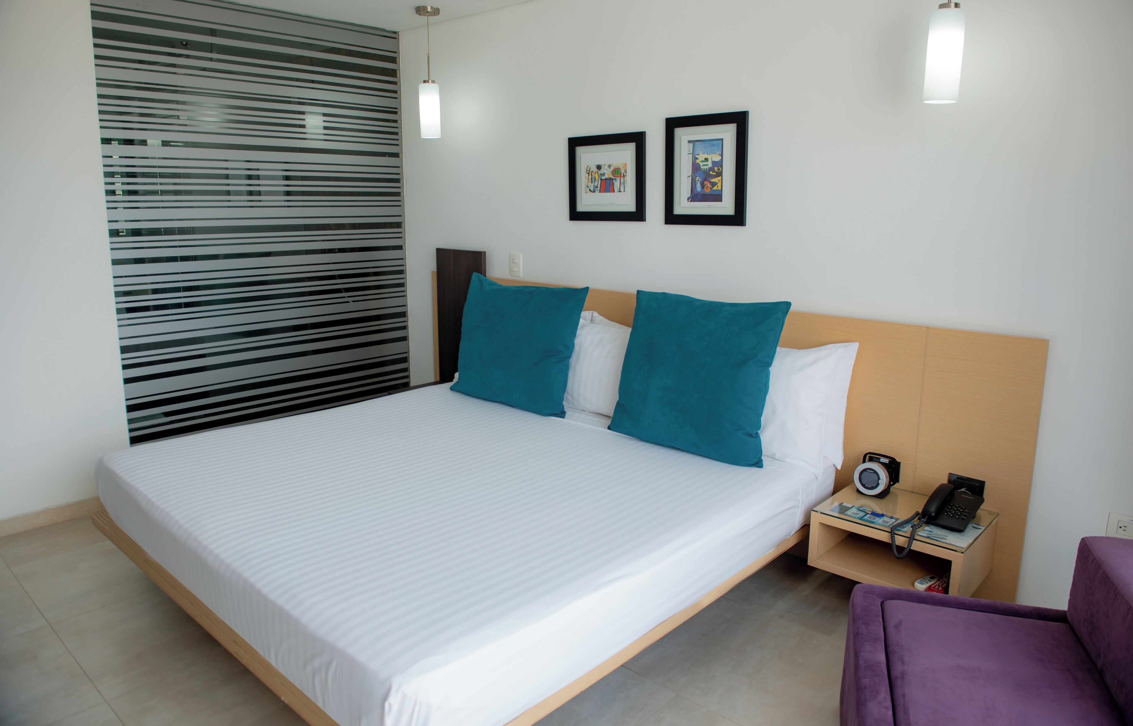 A neatly made bed with white linens and teal pillows, a bedside table with a lamp, and a small purple chair.