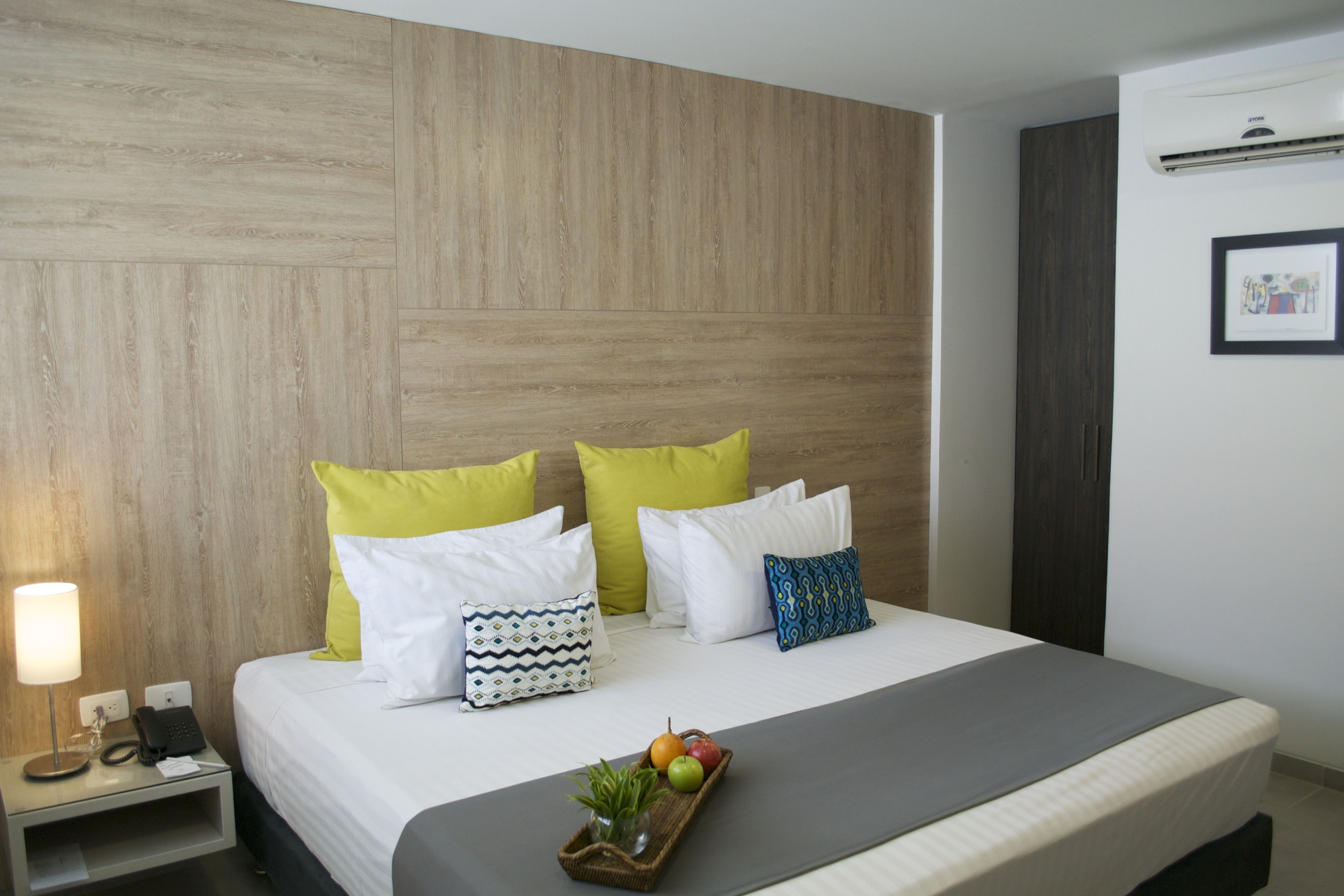 A hotel room with a wooden headboard, a bed with white and patterned pillows, a bedside table with a lamp, a phone, and a tray with fruits.
