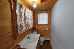 A small, wooden-paneled bathroom with a white sink, toilet, and a window with blinds.