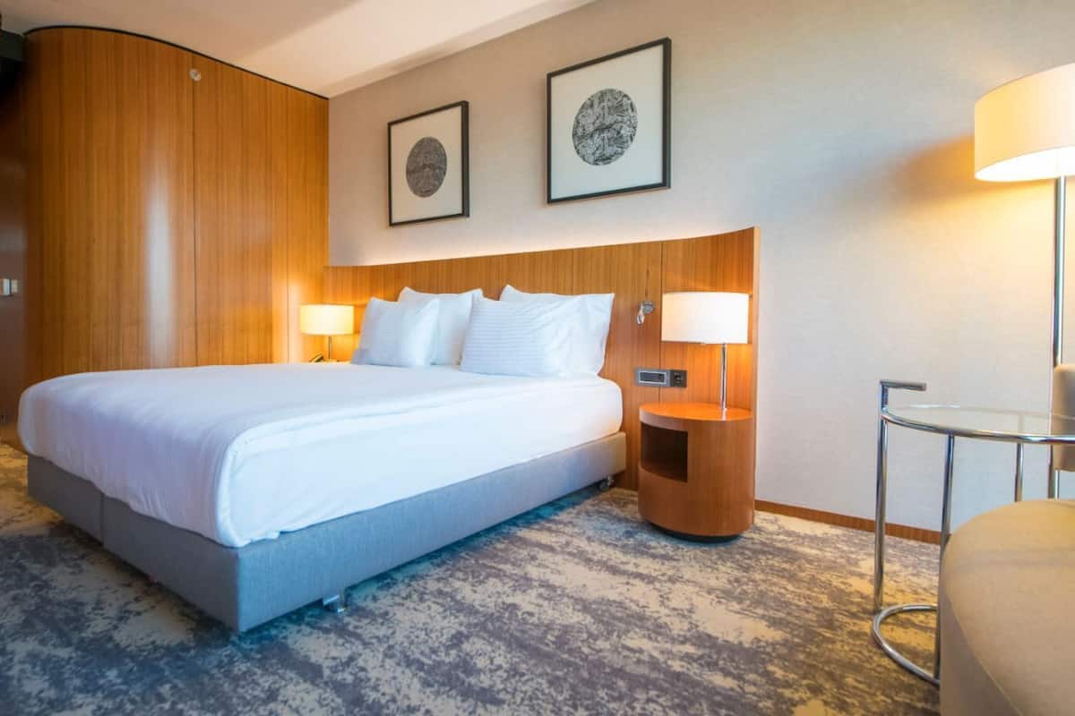 Habitación de hotel con una cama grande, dos lámparas de noche, una mesita de noche redonda de madera, una pequeña mesa redonda y dos cuadros en la pared.
