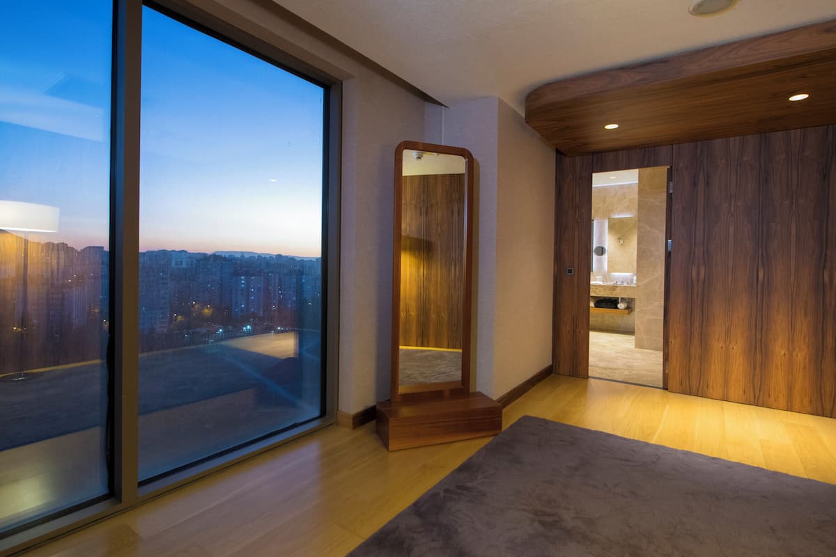 Una habitación de hotel moderna con un ventanal grande, suelo de madera, un espejo y una puerta que da a otra habitación.