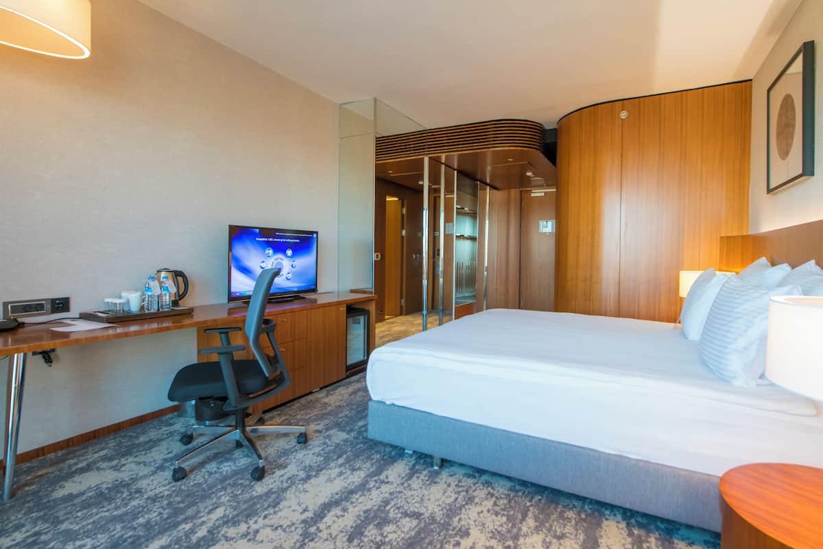 Habitación de hotel con cama, escritorio, silla, televisor y baño visible a través de una puerta de cristal.