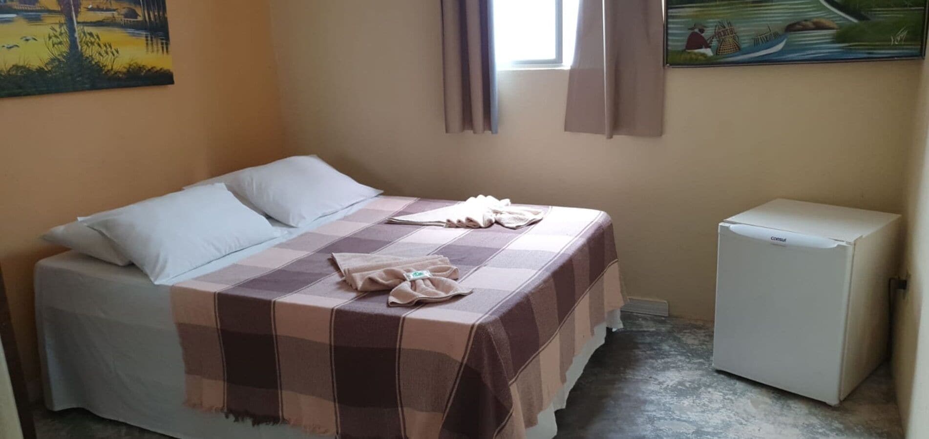 Une chambre à coucher avec un lit, des oreillers, une couverture à carreaux, un petit réfrigérateur et deux tableaux accrochés au mur.
