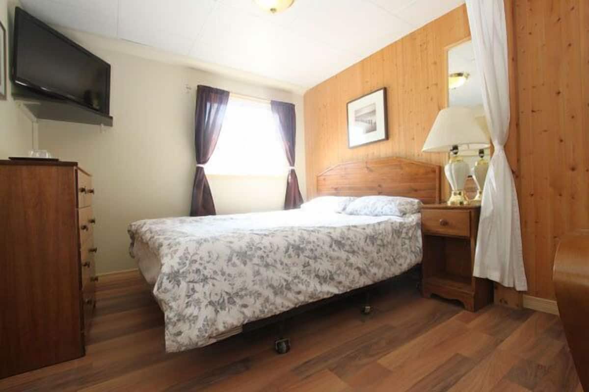 Un dormitorio con cama, cabecera de madera, mesita de noche, lámpara, televisor en la pared y ventana con cortinas.