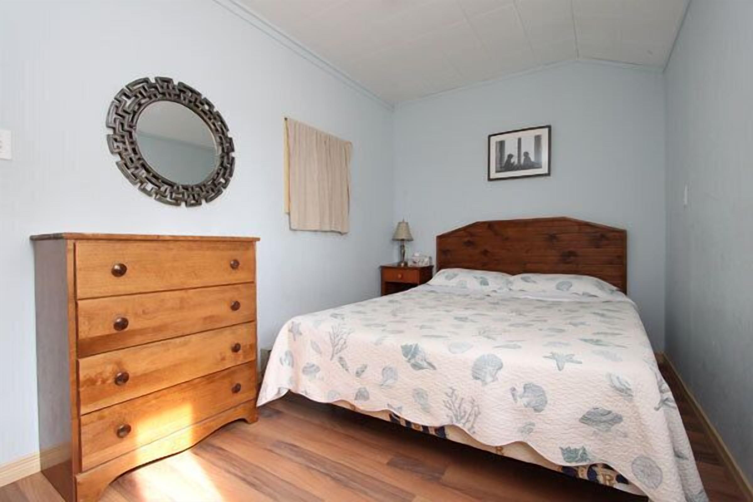 A bedroom with a wooden dresser, a bed with a floral bedspread, a round mirror, a window with a curtain, and a framed picture on the wall.