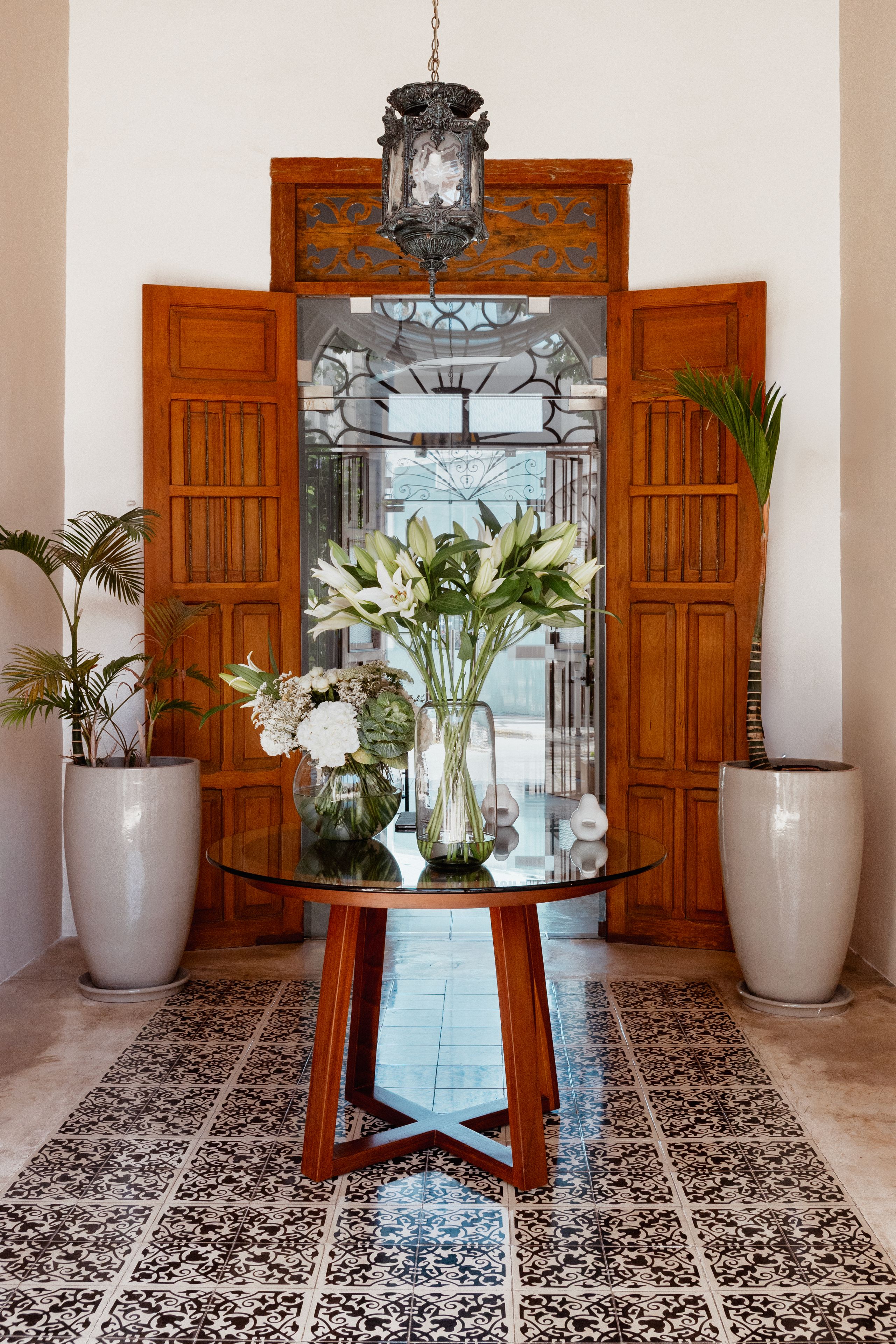 Mesa com tampo de vidro, adornada com um vaso de flores, e acompanhada por dois vasos de plantas, em um quarto com piso de azulejos estampados e portas de madeira.