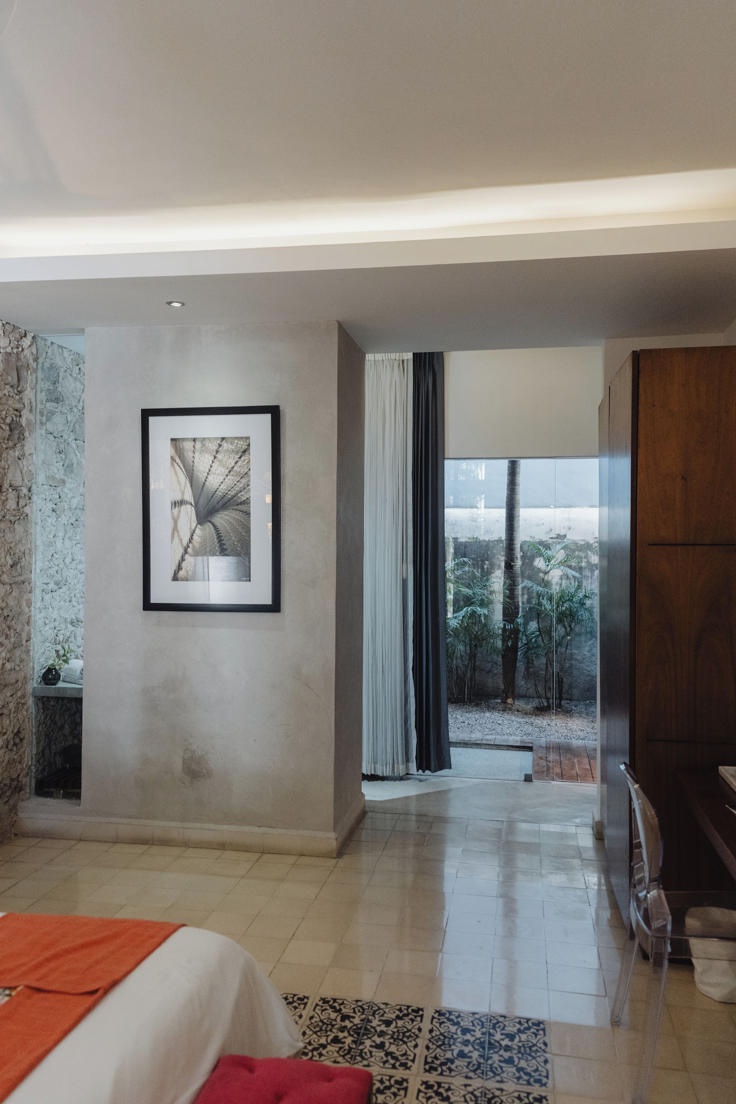 A modern bedroom with a large bed, a framed picture on the wall, and a view of a tropical landscape through a window.
