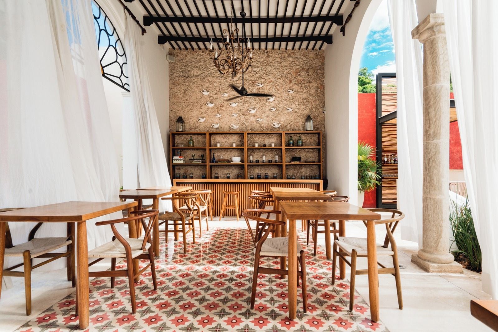 Uma área de refeições com mesas e cadeiras de madeira, piso estampado, parede de pedra com um candelabro e uma estante com diversos objetos.