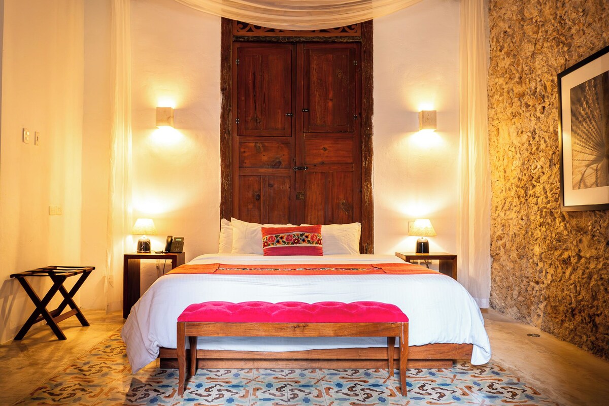 Un dormitorio con una puerta grande de madera, dos lámparas de noche, una cama con una colcha roja y baldosas de suelo con diseño.