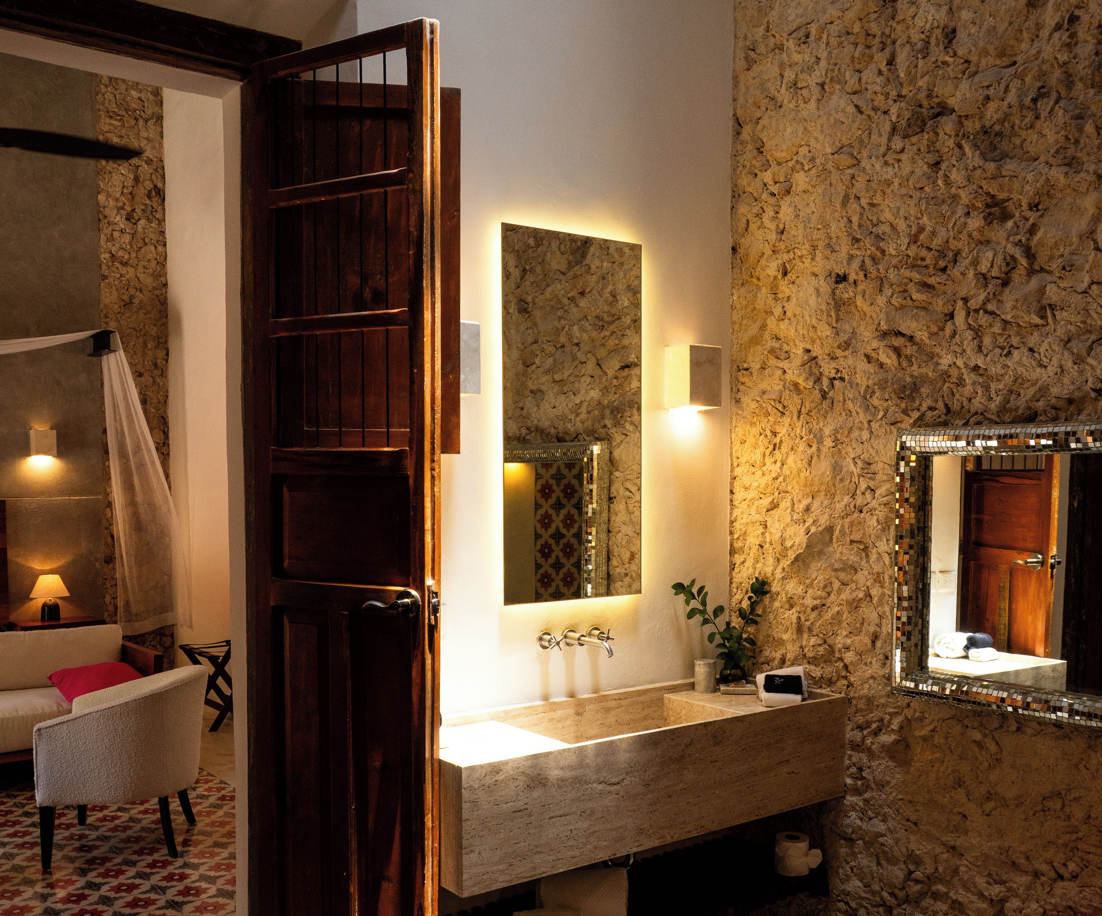 A bathroom with a stone wall, a wooden door, a sink with a mirror, and a towel rack.