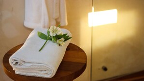 Una toalla blanca doblada con un pequeño arreglo floral sobre un taburete de madera en una habitación con pared blanca y un interruptor de luz a la vista.