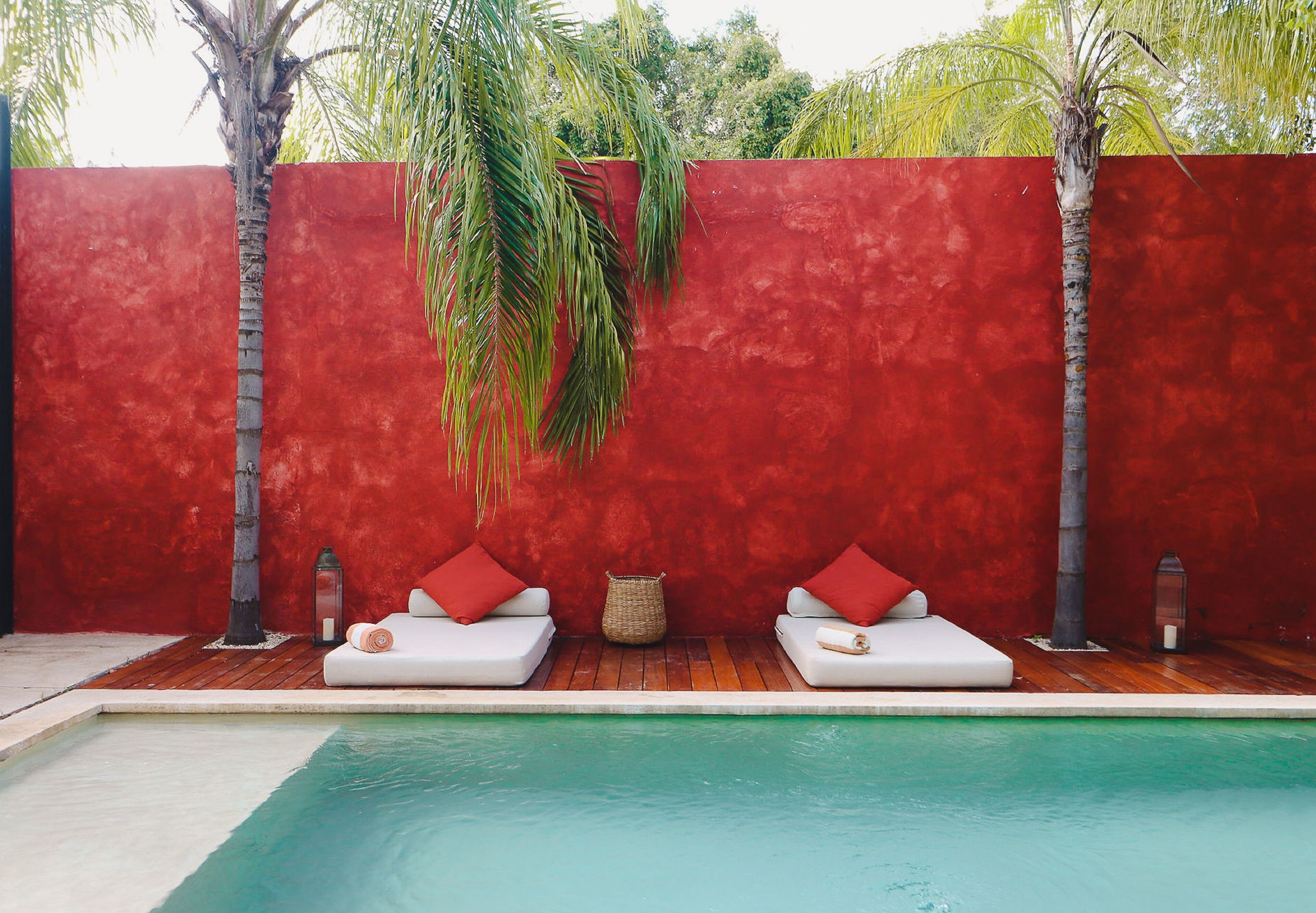 Un área junto a la piscina con dos sillones blancos, cojines rojos, una cesta tejida y una pared roja.