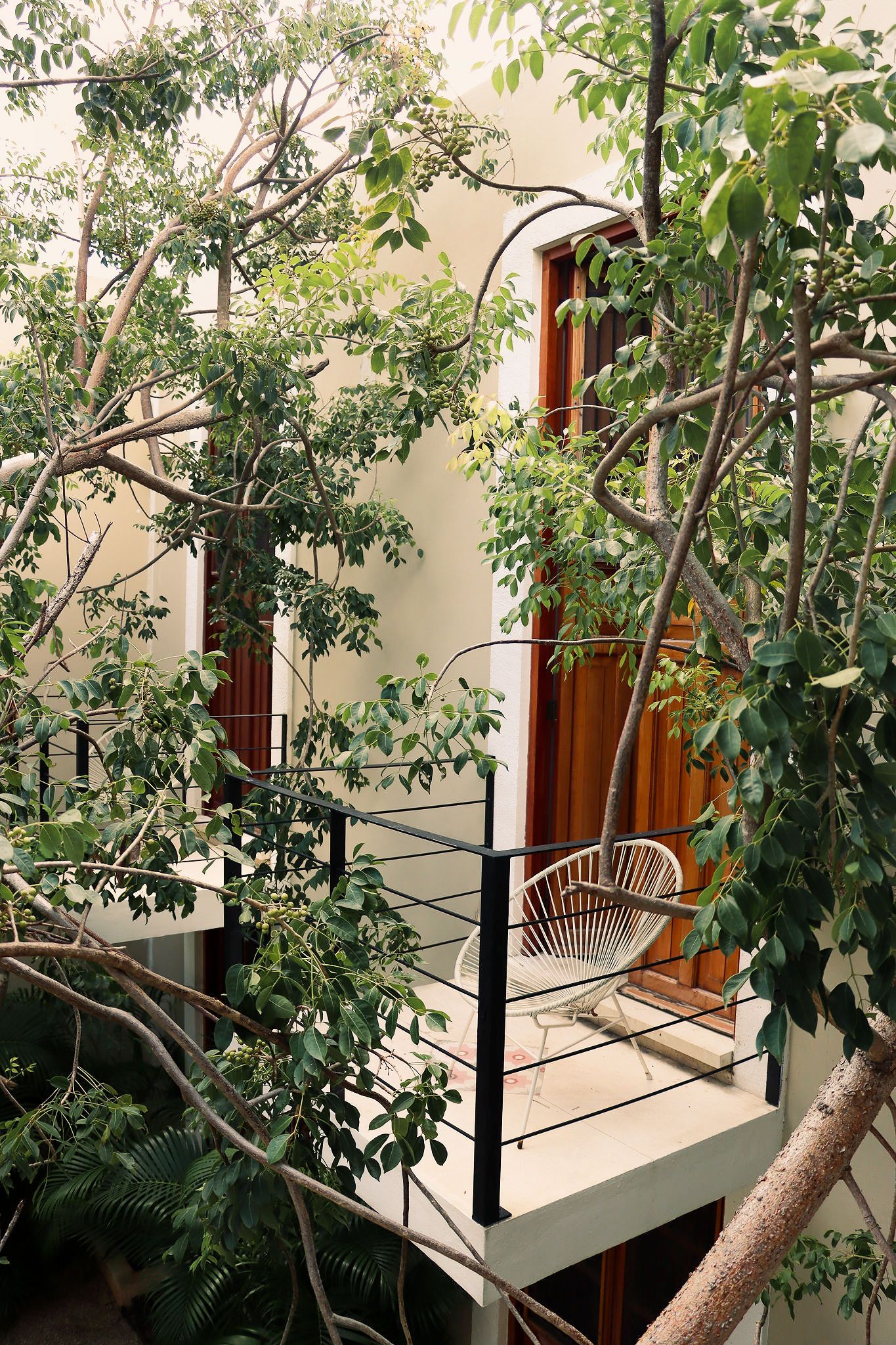 A balcony with a white chair, surrounded by lush greenery and a building with a wooden door.