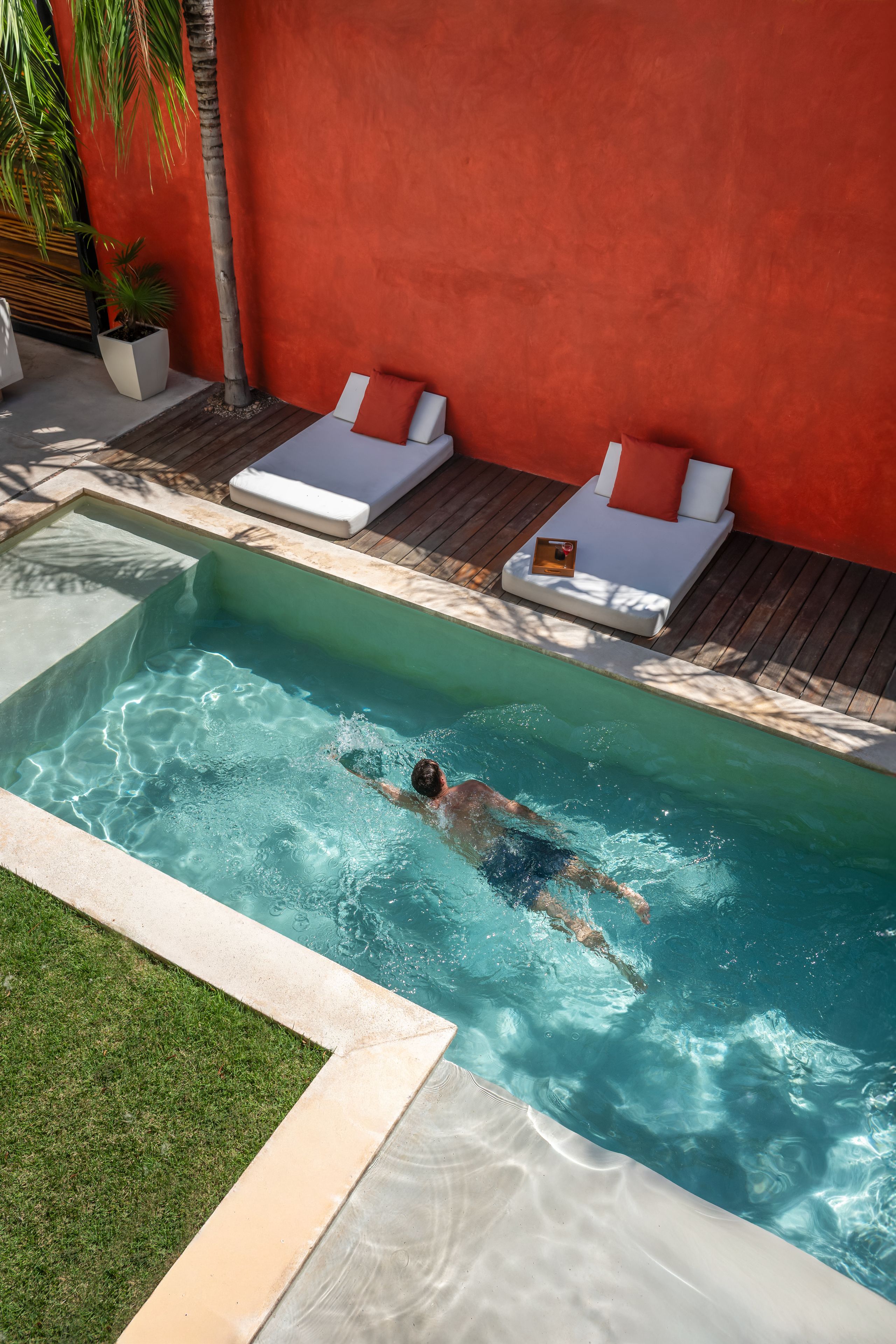 A swimming pool with a person swimming, lounge chairs, and a red wall.