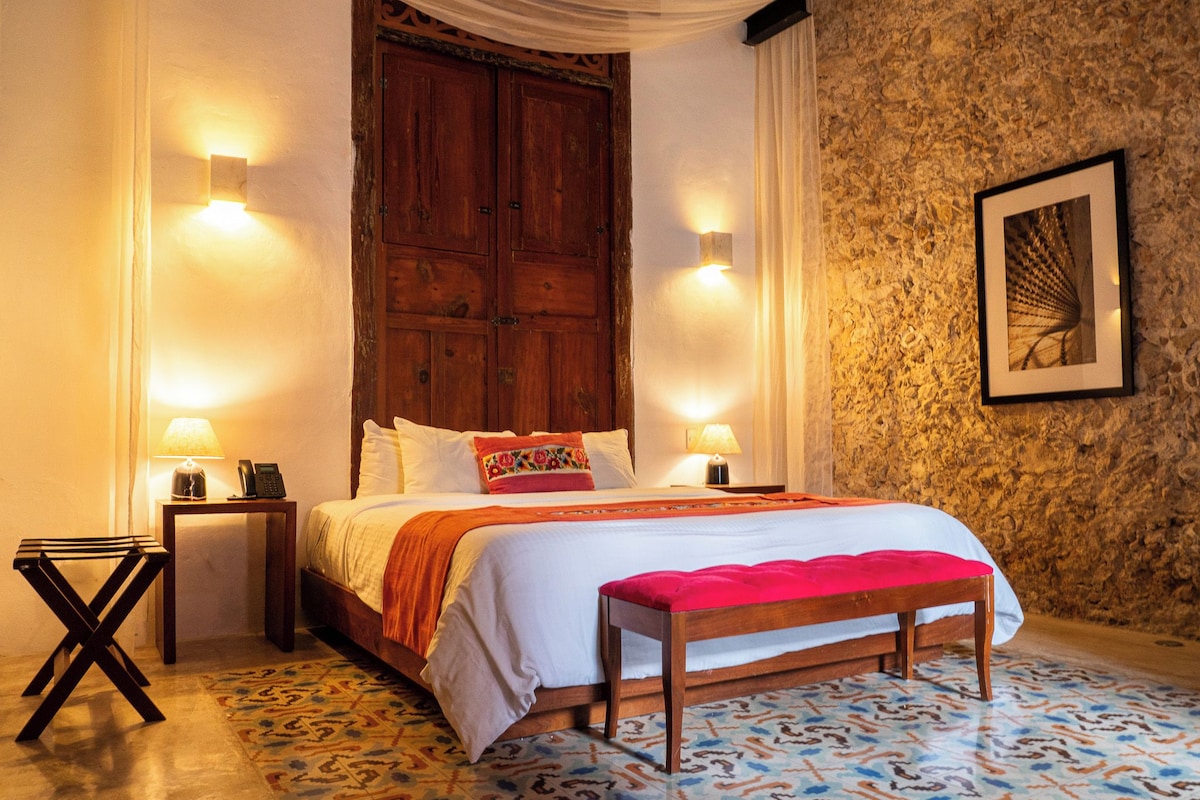 A bedroom with a large wooden door, two bedside lamps, a bed with a red blanket, a bench, and a patterned floor.