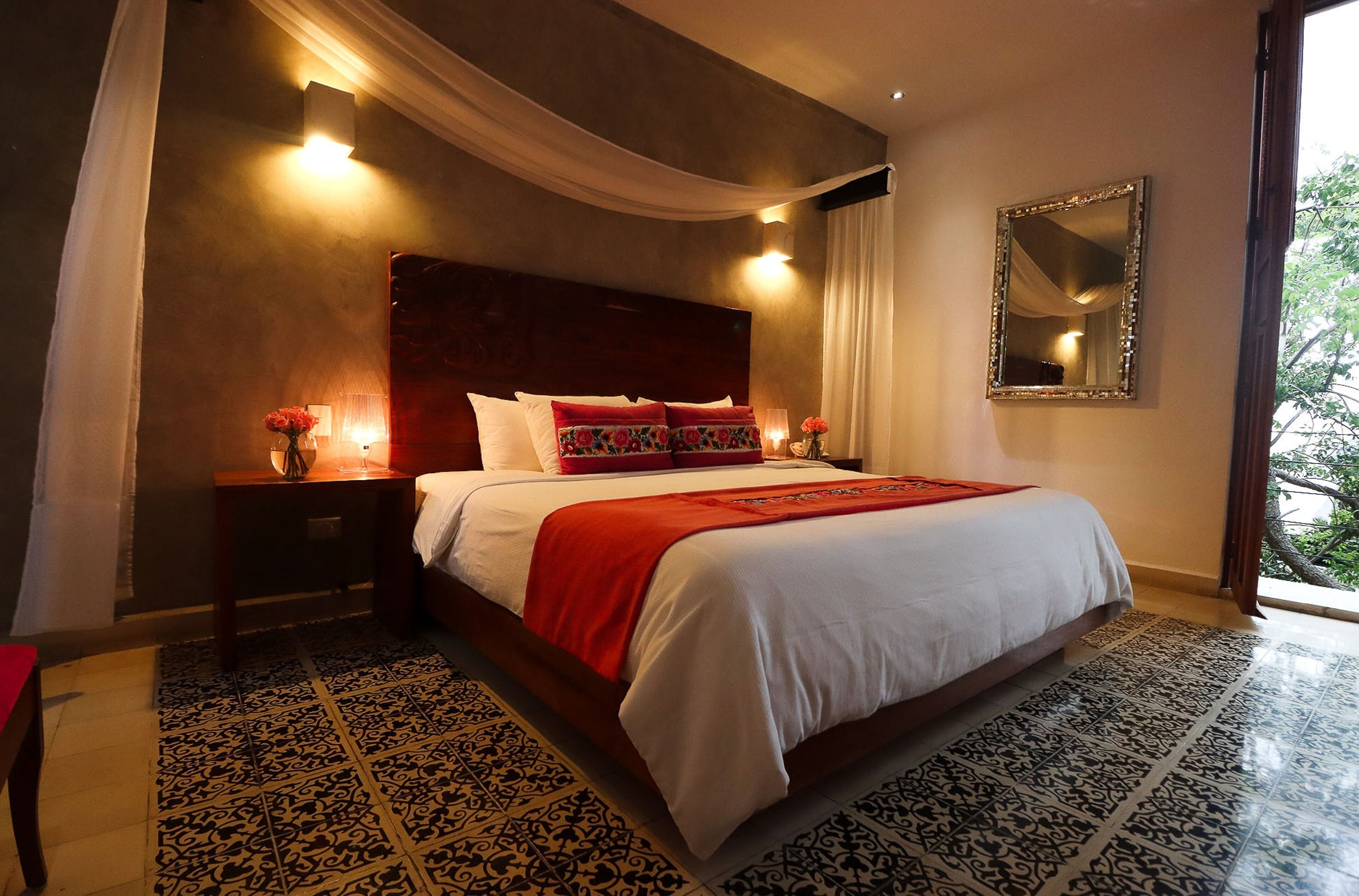 Une chambre d’hôtel avec un grand lit, une table de chevet ornée d’un vase de fleurs, un miroir au mur et une vue sur la verdure à l’extérieur.