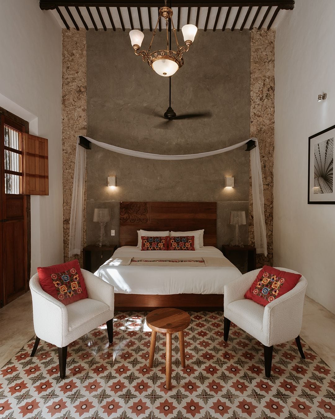 A bedroom with a bed, two chairs, a small table, a ceiling fan, and a wall-mounted light fixture.